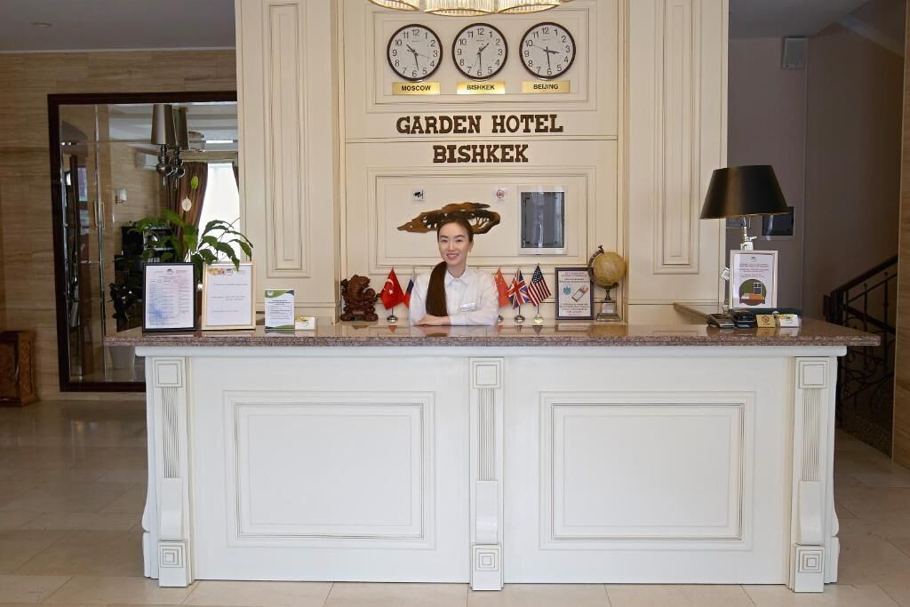 A reception desk with clocks displaying Moscow, Bishkek, and Beijing times, a sign for Garden Hotel Bishkek, and various flags.