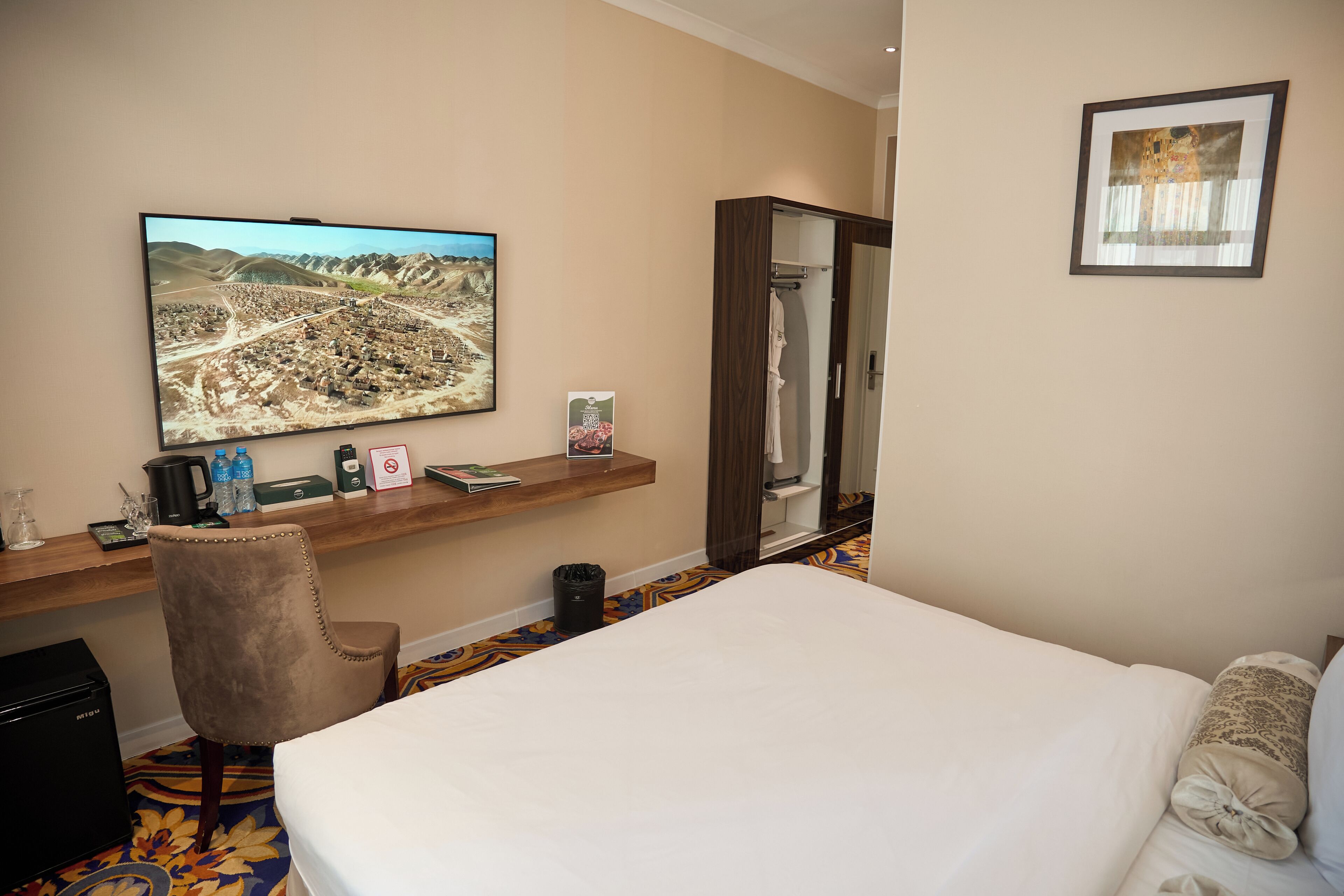 A hotel room with a bed, a desk with a flat-screen TV, a chair, a wardrobe, and a framed picture on the wall.