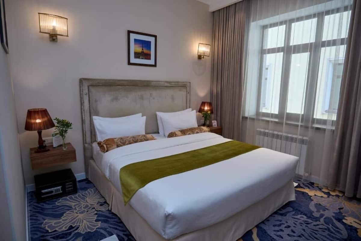 Una habitación de hotel con una cama grande, dos lámparas de noche, un cuadro en la pared y una ventana con cortinas.