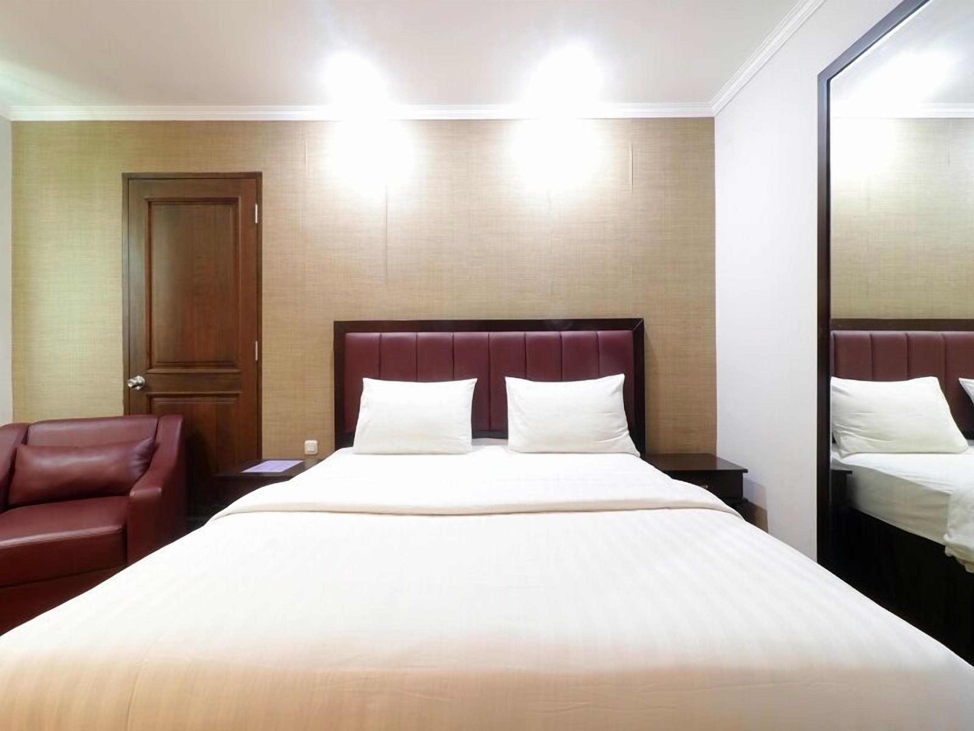 A hotel room with a large bed, a wooden door, a red leather chair, and a mirror on the wall.