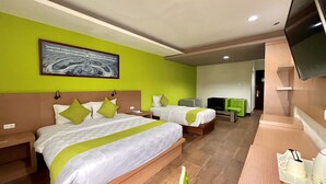 Habitación de hotel con dos camas, televisión, escritorio con cafetera y pared verde con una cita motivacional.