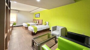 Une chambre d’hôtel avec deux lits, une table basse en verre, un téléviseur et un coin salon avec des fauteuils verts.
