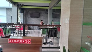 Un espace d’accueil d’hôtel avec un comptoir de réception en bois portant l’inscription « CONCIERGE », un coin salon avec des tables et des chaises, et une plante en pot ornée de différentes fleurs.