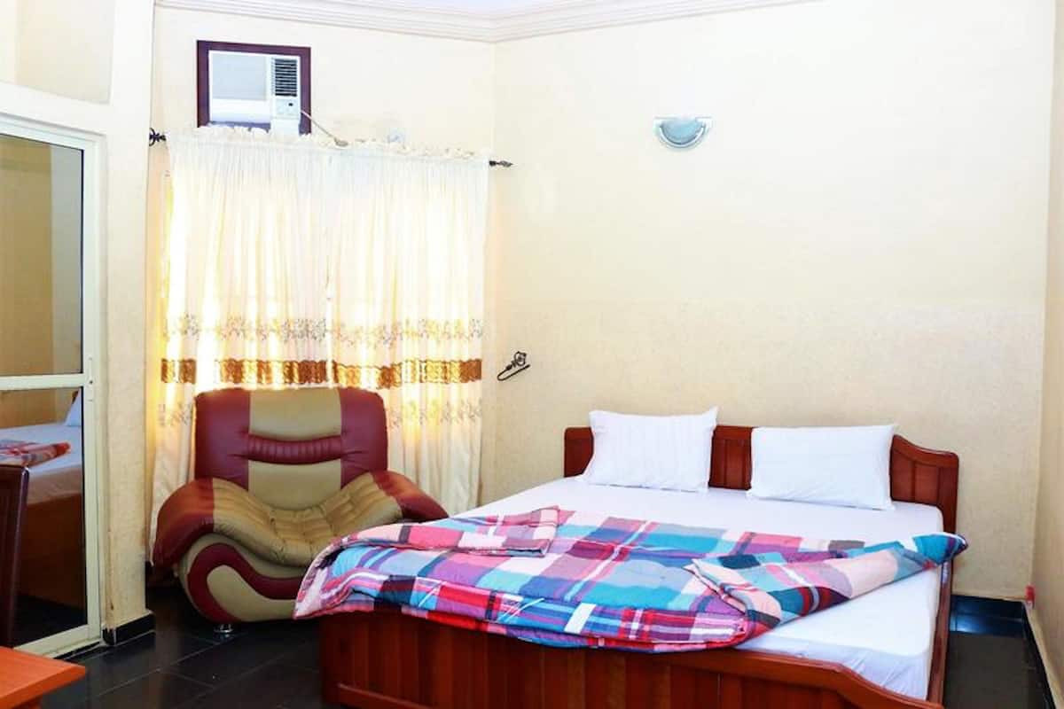 A bedroom with a bed, a chair, a window with curtains, and a thermostat on the wall.
