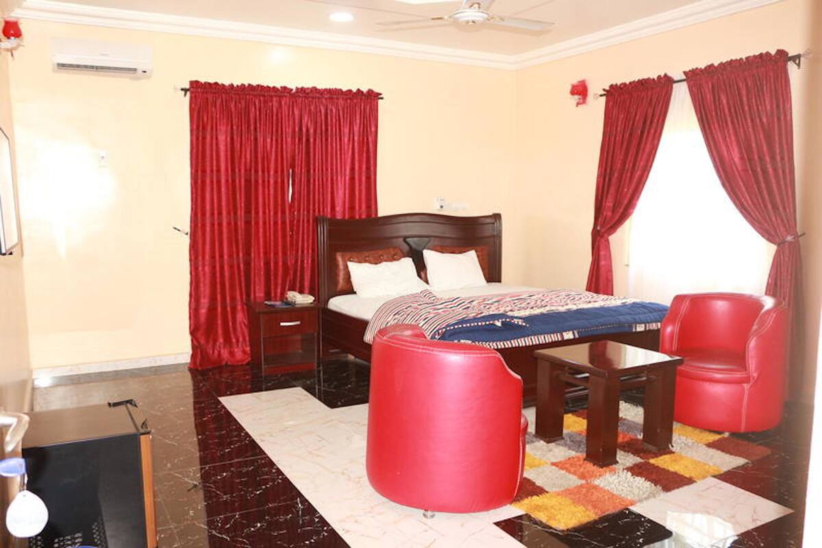 A hotel room with a bed, a red ottoman, a red chair, a small table, and a window with red curtains.