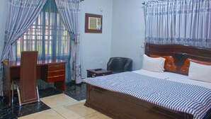 Un dormitorio con cama, escritorio y silla. Se ve una ventana con cortinas, un aire acondicionado de pared y un cuadro enmarcado.