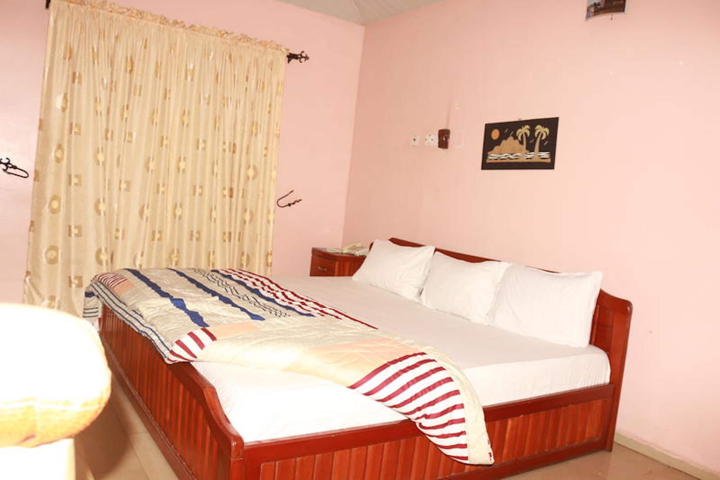 A bedroom with a wooden bed, white bedding, a patterned blanket, a window with curtains, and a wall-mounted picture.