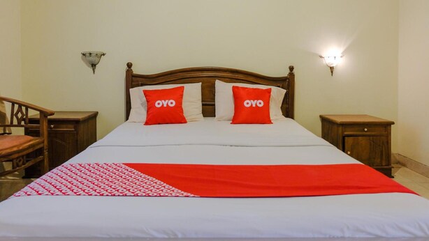 Una cama bien tendida con una colcha blanca y roja, dos almohadas rojas con la marca "OYO" y un cabecero de madera.