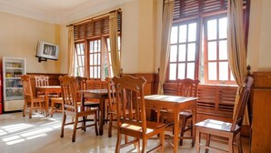 Un comedor con mesas y sillas de madera, ventanas amplias con cortinas, un televisor en la pared y un refrigerador en la esquina.