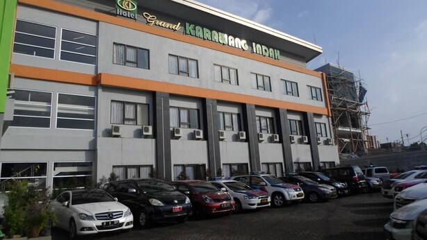 Un hotel llamado Grand Karawang Indah con una fachada moderna, varios equipos de aire acondicionado y un estacionamiento con diferentes automóviles.