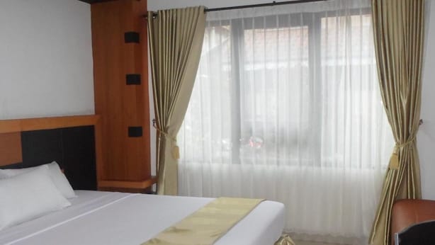A hotel room with a bed, a chair, a wooden headboard, and an air conditioning unit. - Hotel Grand Karawang Indah (Karawang)