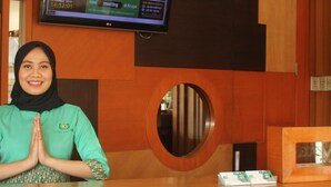 Un mostrador de recepción de hotel con un televisor mostrando un anuncio de reunión, una zona de recepción de madera y un espejo circular.