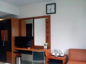 A hotel room with a wooden desk, a television, a large mirror, a sofa, and a clock on the wall.