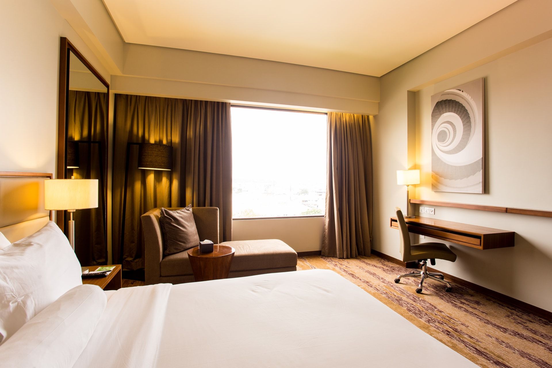 Une chambre d’hôtel comprenant un lit, un bureau, une chaise, une lampe, un miroir et une fenêtre avec des rideaux.