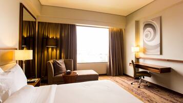 Une chambre d’hôtel comprenant un lit, un bureau, une chaise, une lampe, un miroir et une fenêtre avec des rideaux.