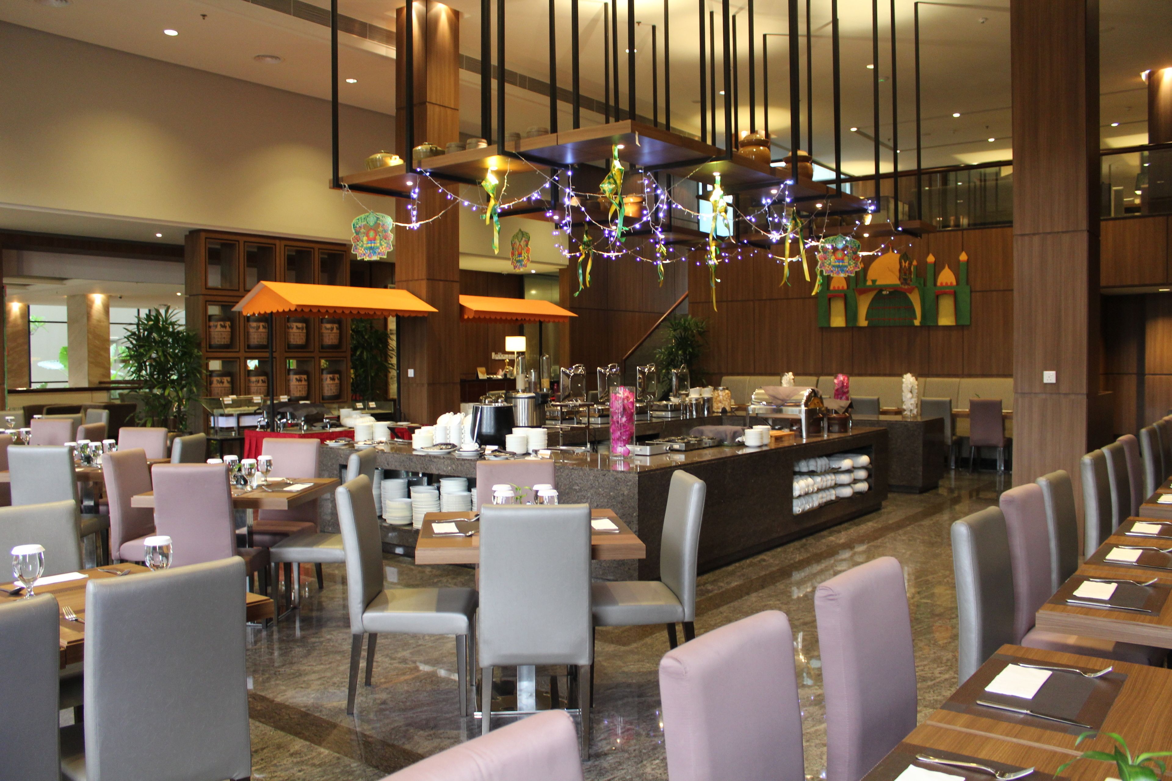 A hotel restaurant with a long counter, multiple tables, and chairs. There are decorative lights hanging from the ceiling.