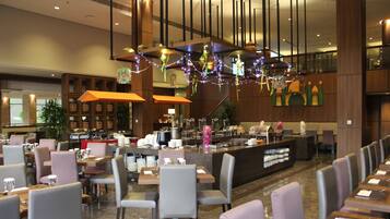 A hotel restaurant with a long counter, multiple tables, and chairs. There are decorative lights hanging from the ceiling.