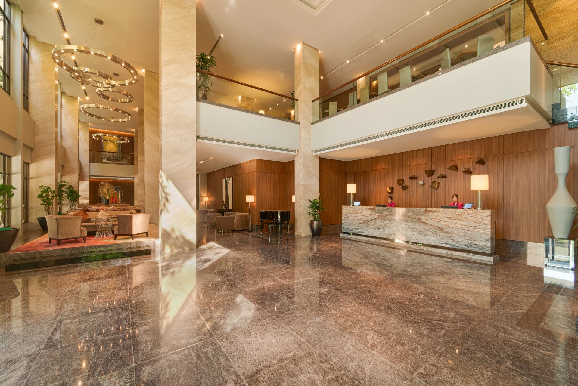 A spacious lobby with a marble reception desk, seating areas, and a mezzanine level with glass railings.