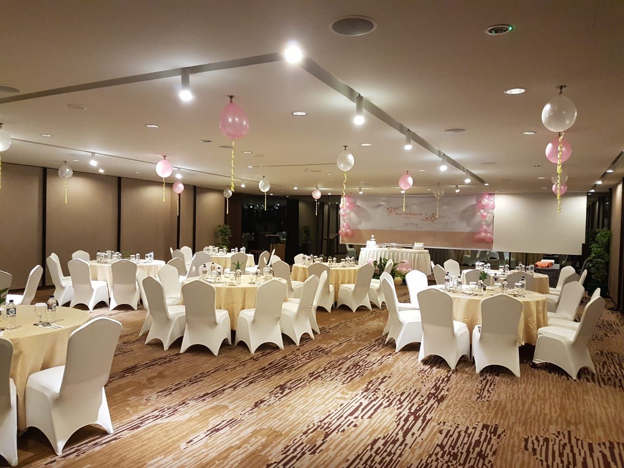 Un salón de banquetes con mesas redondas, sillas blancas y piso alfombrado.