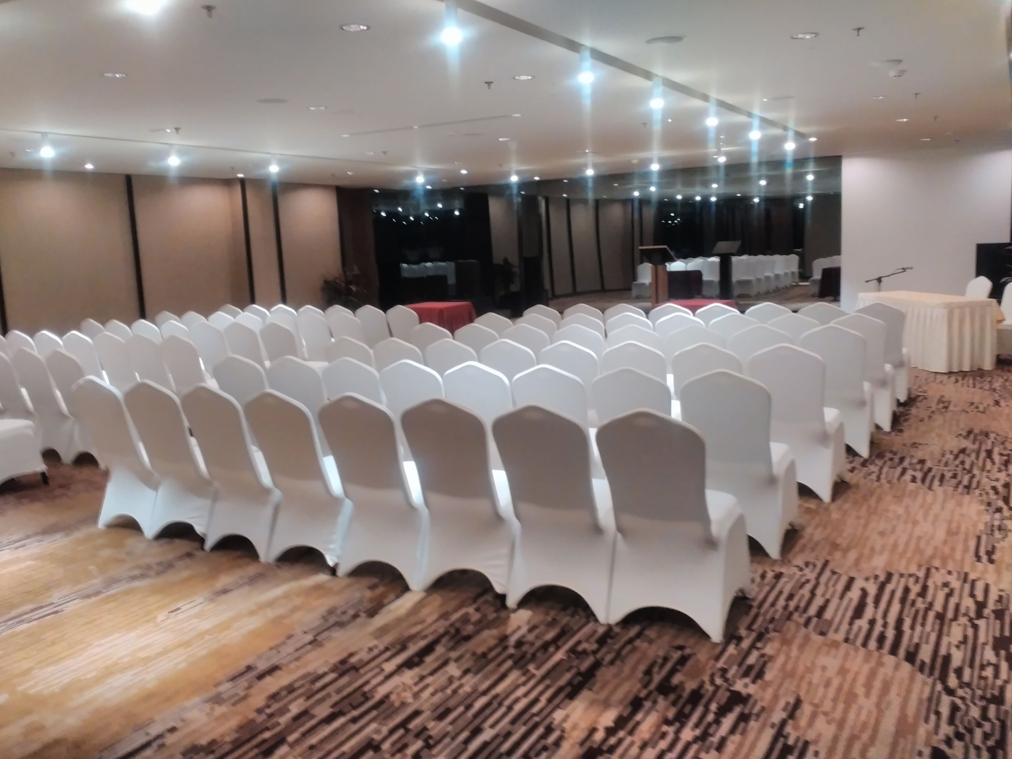 A conference room with rows of white chairs, a stage with a podium, and a carpeted floor.