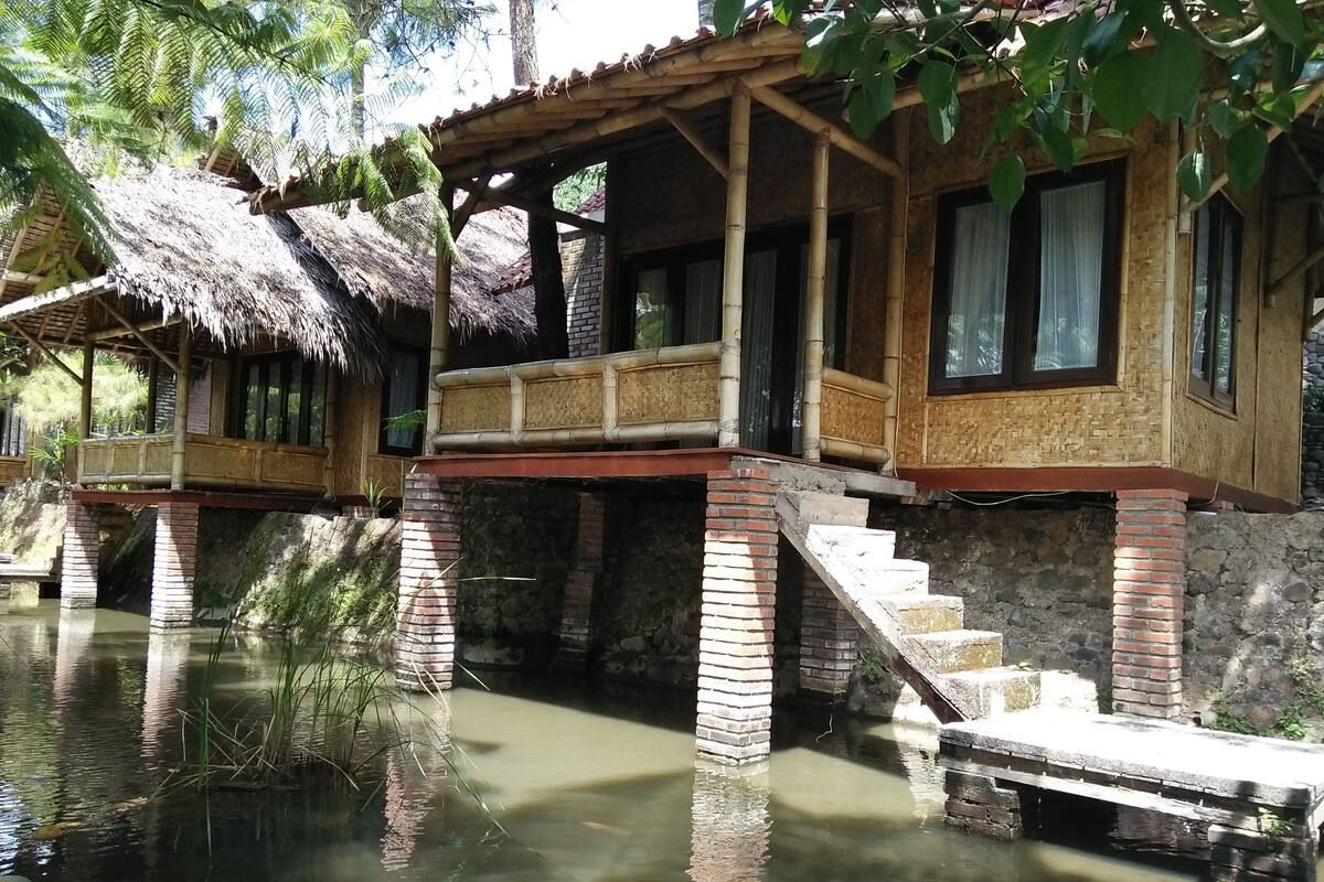 Uma casa tradicional de madeira com telhado de palha, estrutura de bambu e escadaria de pedra que leva a um deck de madeira sobre a água.
