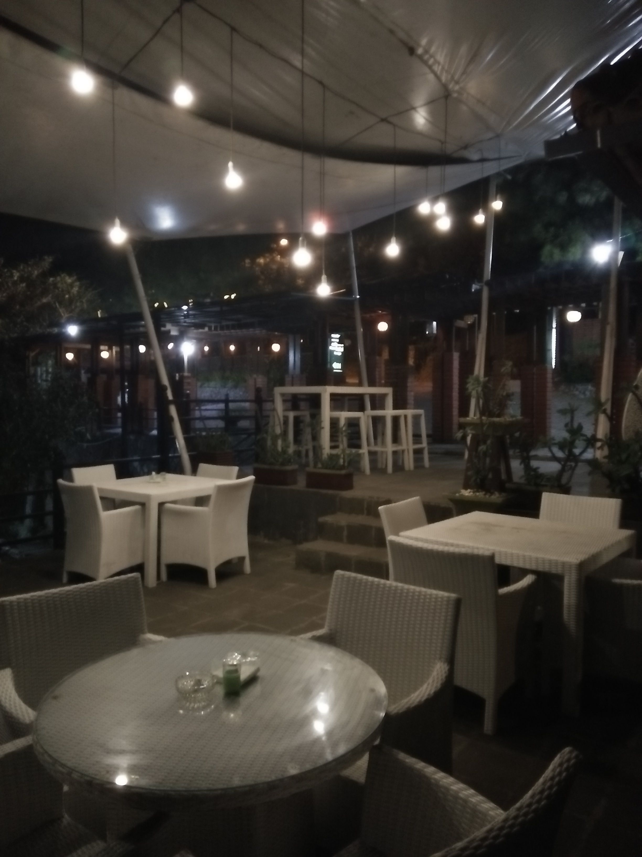 An outdoor dining area with round tables and chairs, overhead lighting, and potted plants.
