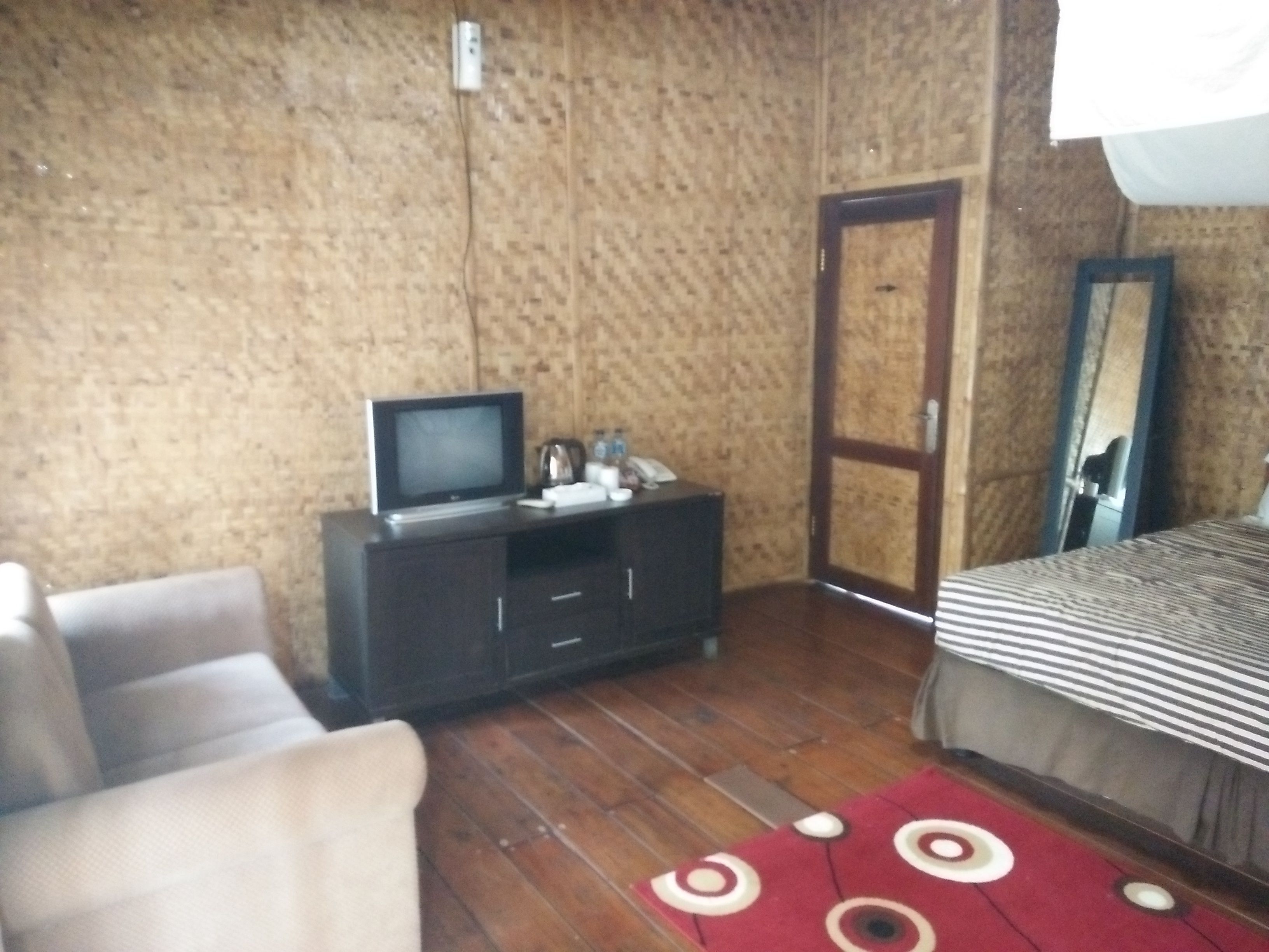 A room with a wooden floor, a bed with a striped bedspread, a dark-colored TV stand with a television, a mirror, and a red rug with circular patterns.