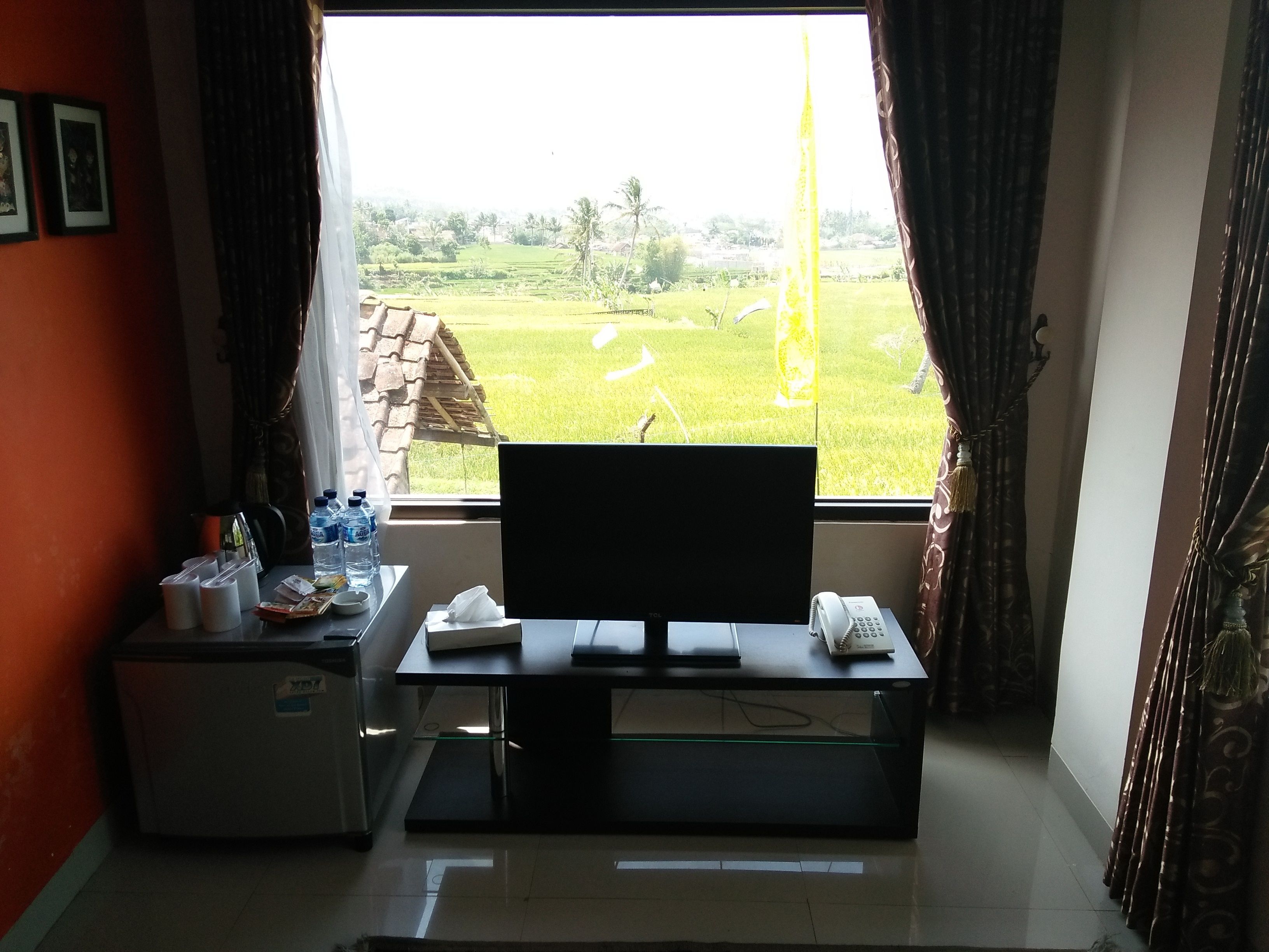 Una habitación con escritorio, silla, refrigerador pequeño y ventana con cortinas.