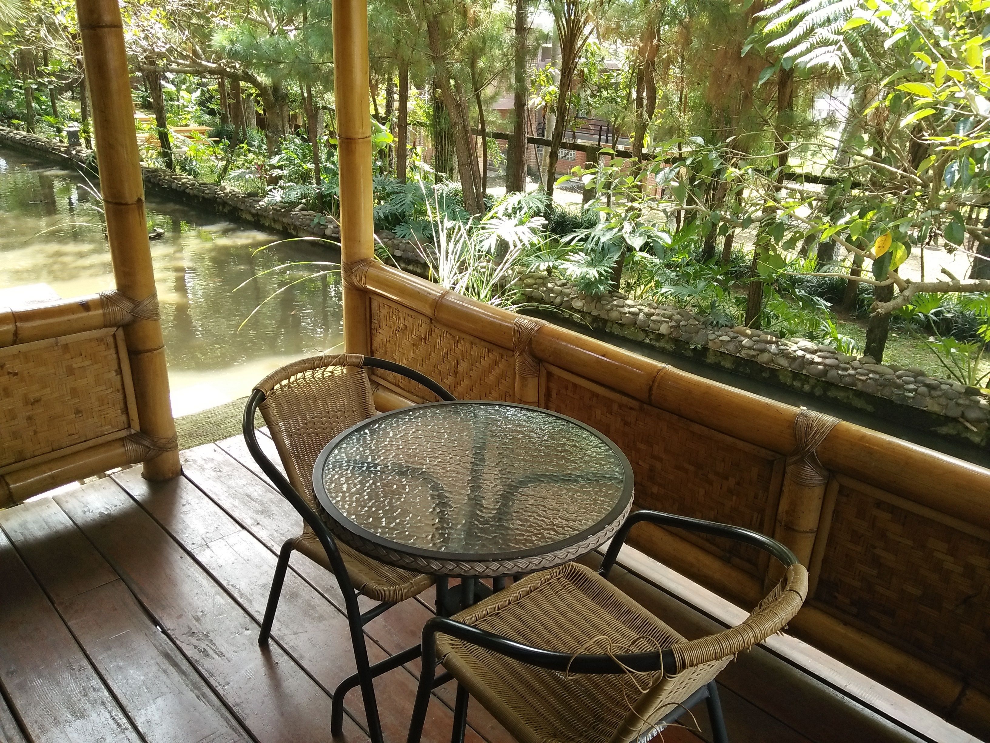 A bamboo-framed balcony with a glass table and wicker chairs overlooking a pond and lush greenery.
