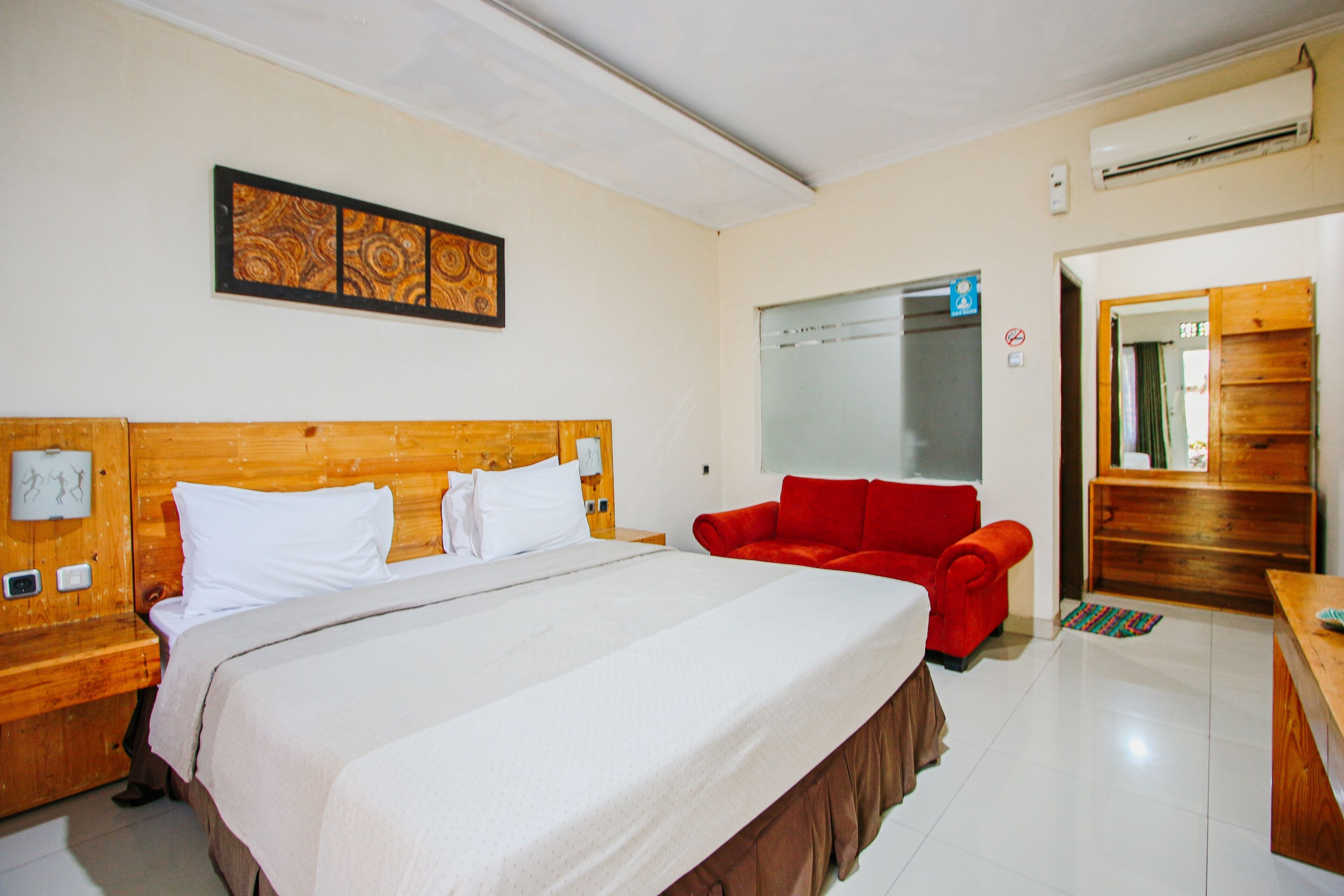 Una habitación de hotel con una cama grande, un sillón rojo, un aparador de madera y un aire acondicionado instalado en la pared.
