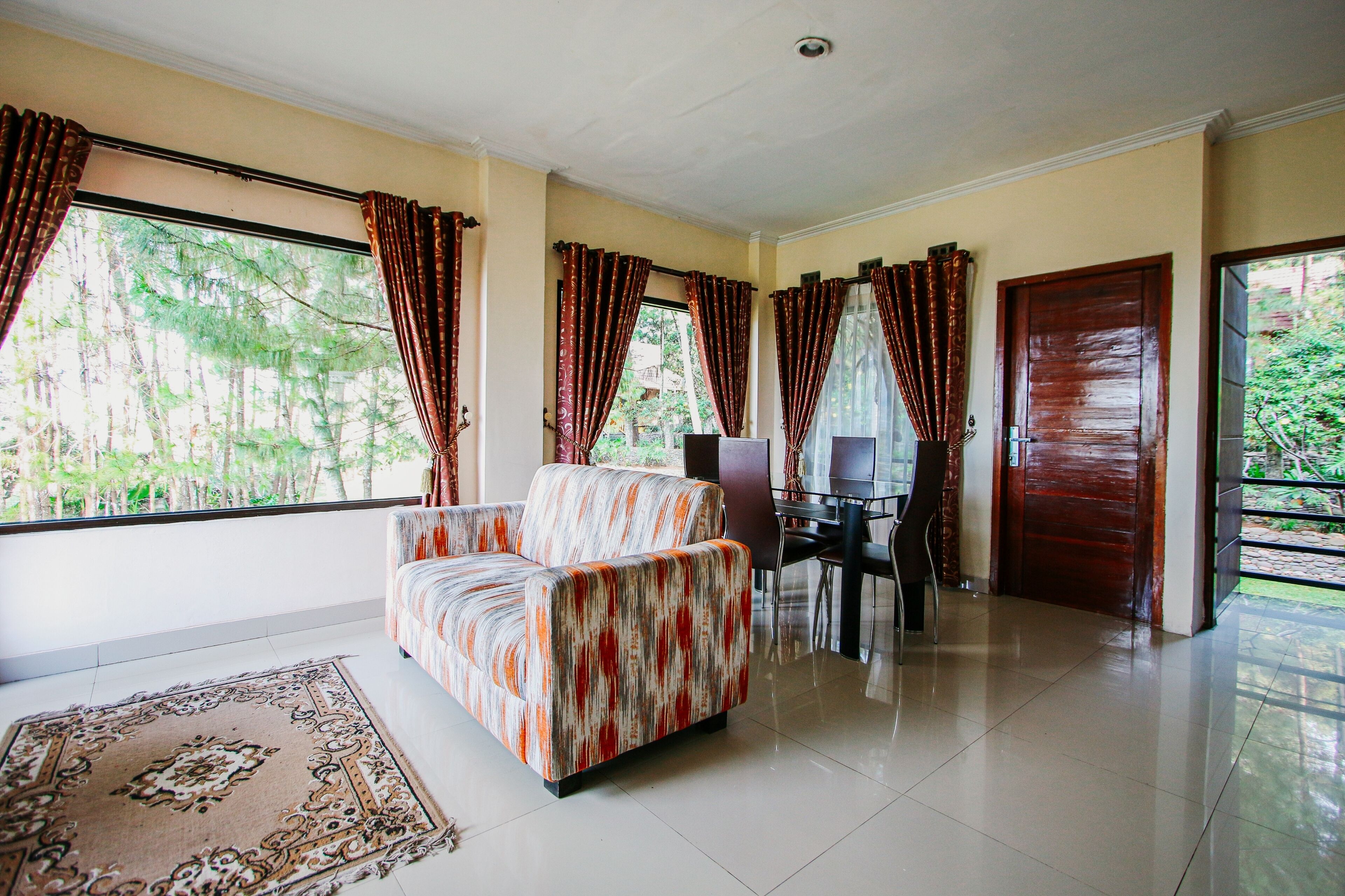 A spacious living room with a sofa, a dining table, and chairs. There are large windows with curtains, and a door leading outside.