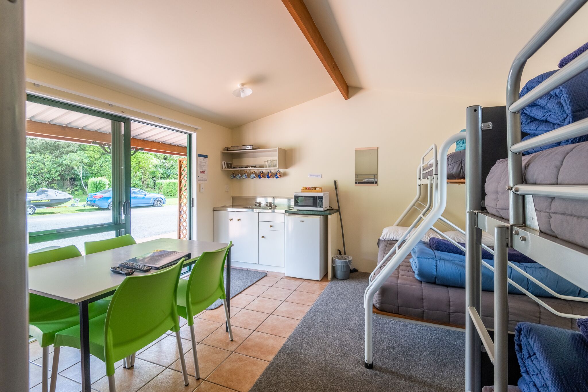A room with bunk beds, a dining table with chairs, a kitchen area, and a view through a large window.