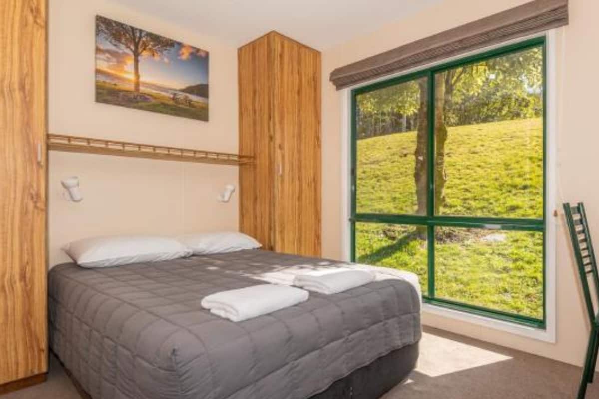 Une chambre avec une grande fenêtre, un lit recouvert d’une couette grise, des lambris en bois sur les murs et un tableau représentant un paysage.