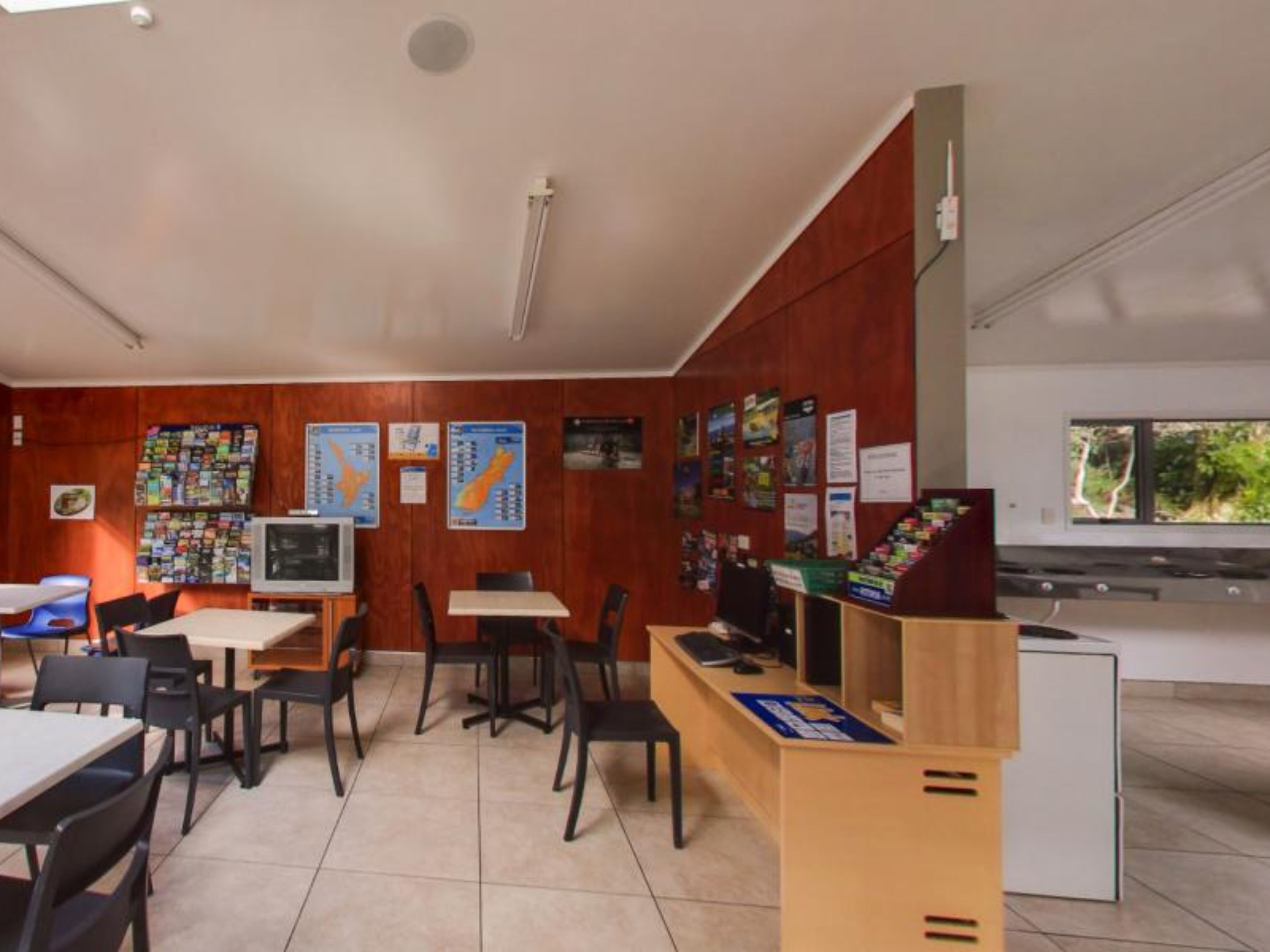 A modern room with wooden panel walls, a tiled floor, and a small TV. There are tables and chairs, a wooden counter, and various posters on the walls.