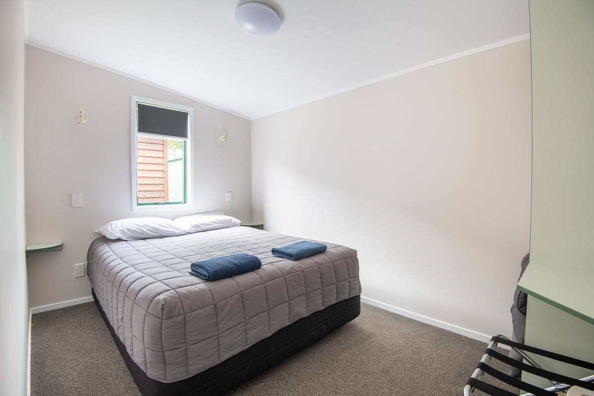 Une petite chambre à coucher, bien rangée, avec un lit simple, une fenêtre et un fauteuil vert.