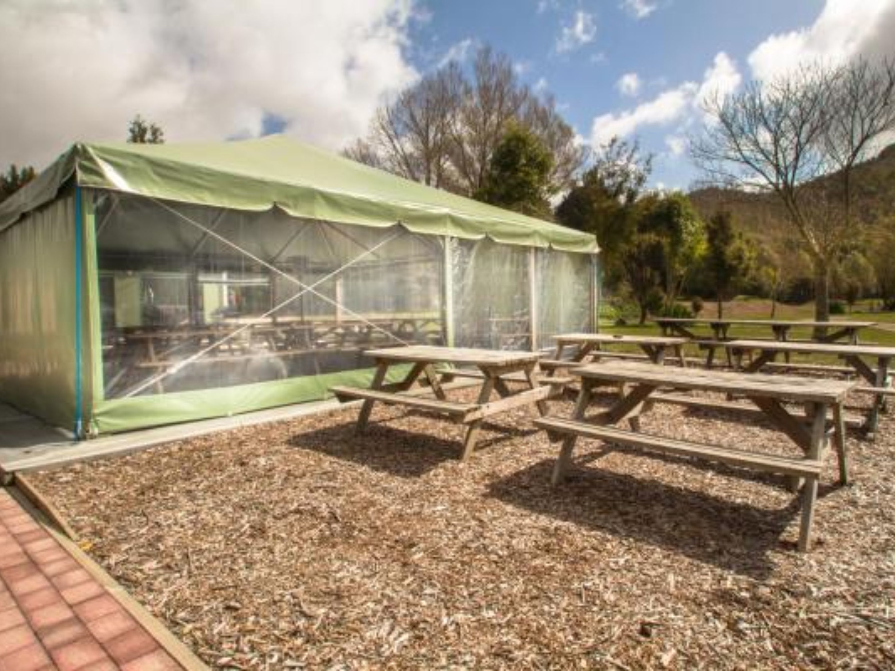 A greenhouse structure with a transparent roof, surrounded by wooden picnic tables on a gravel surface.