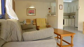 Ein Wohnzimmer mit Sofa, Sessel und Holztisch. Im Hintergrund ist eine Küchenzeile mit Schränken und einem Kühlschrank zu sehen.