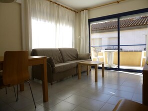 A living room with a sofa, a wooden table, and a chair. There is a large window with curtains and a view of a balcony with a tiled roof.