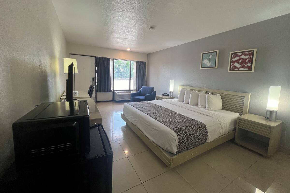A hotel room with a large bed, a desk with a computer, a chair, a desk lamp, and a bedside table with a lamp.
