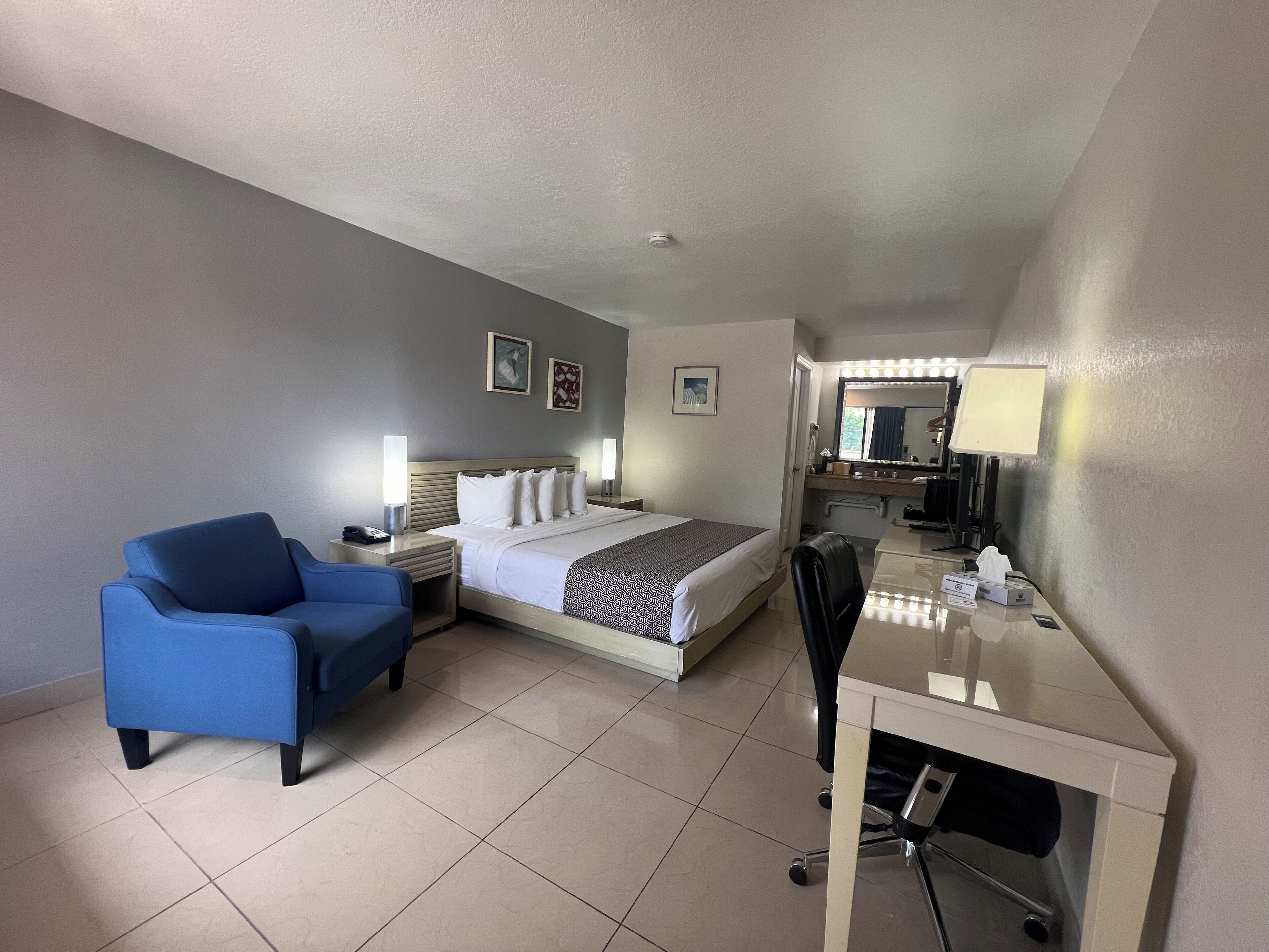 Ein Hotelzimmer mit Bett, Schreibtisch, Stuhl, Schreibtischlampe, Spiegel und Nachttisch mit Lampe.