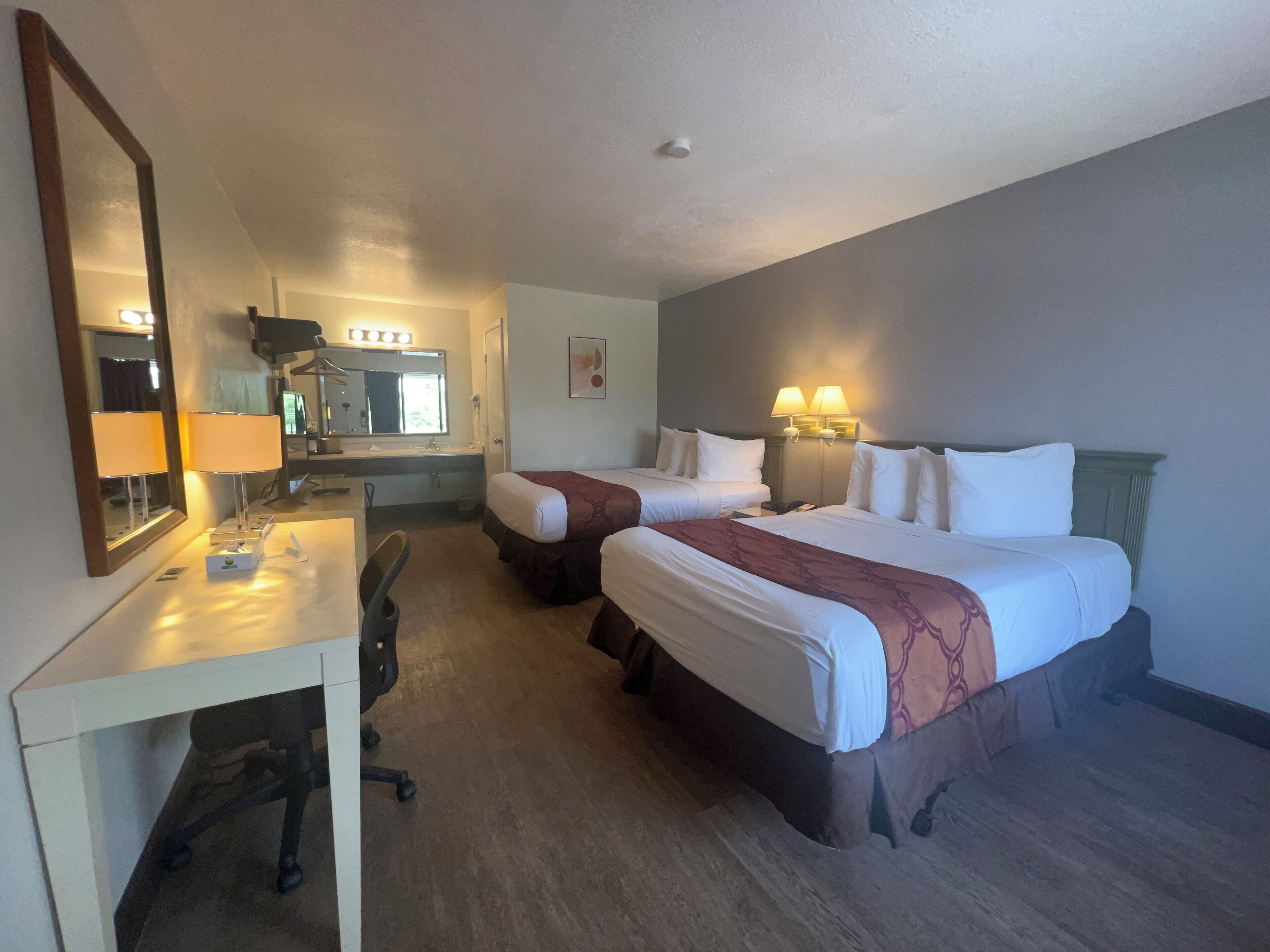 A hotel room with two beds, a desk with a chair, a mirror, and a bathroom visible through an open door.