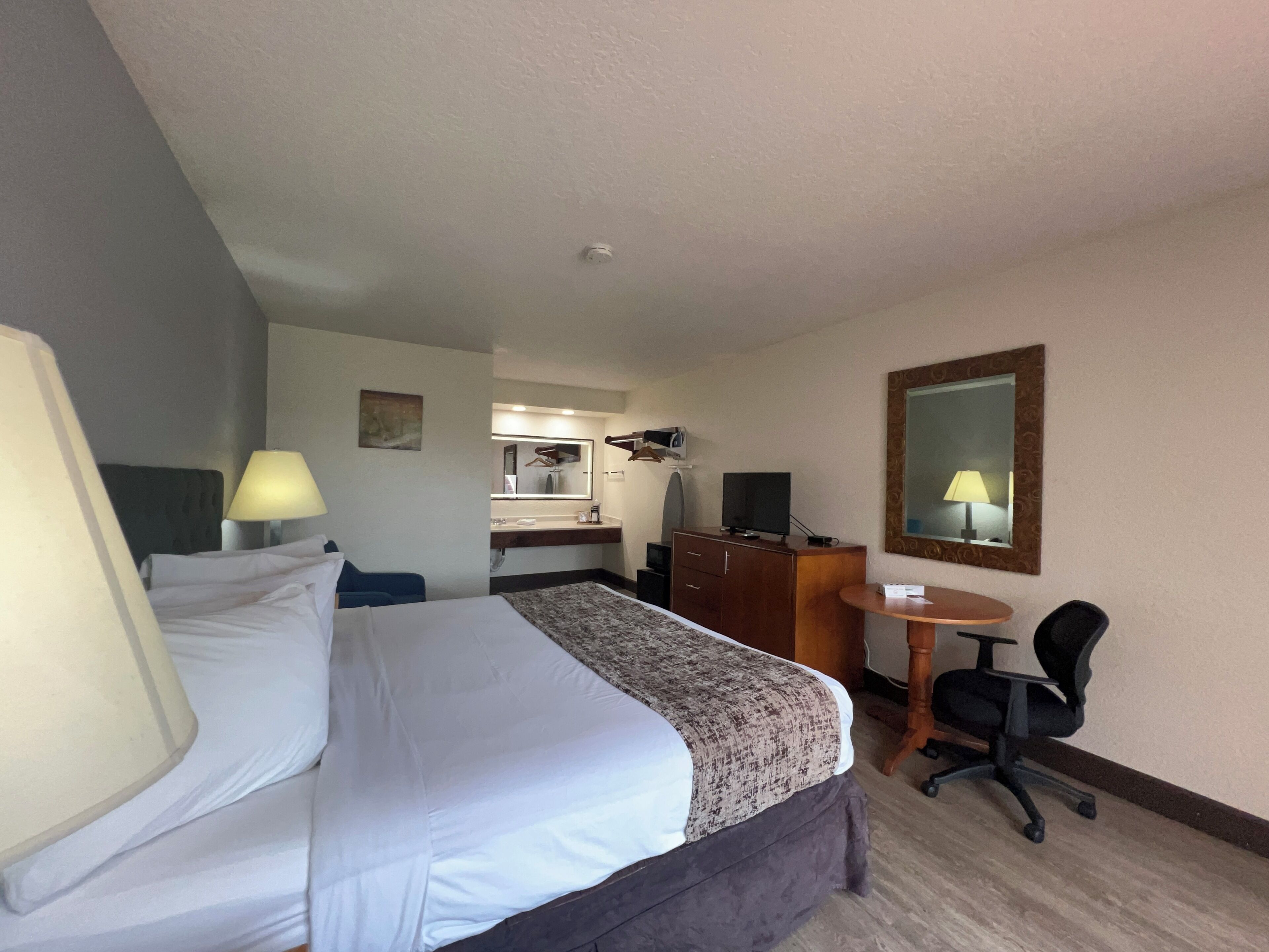 A hotel room with a bed, a desk, a chair, a mirror, and a small table.