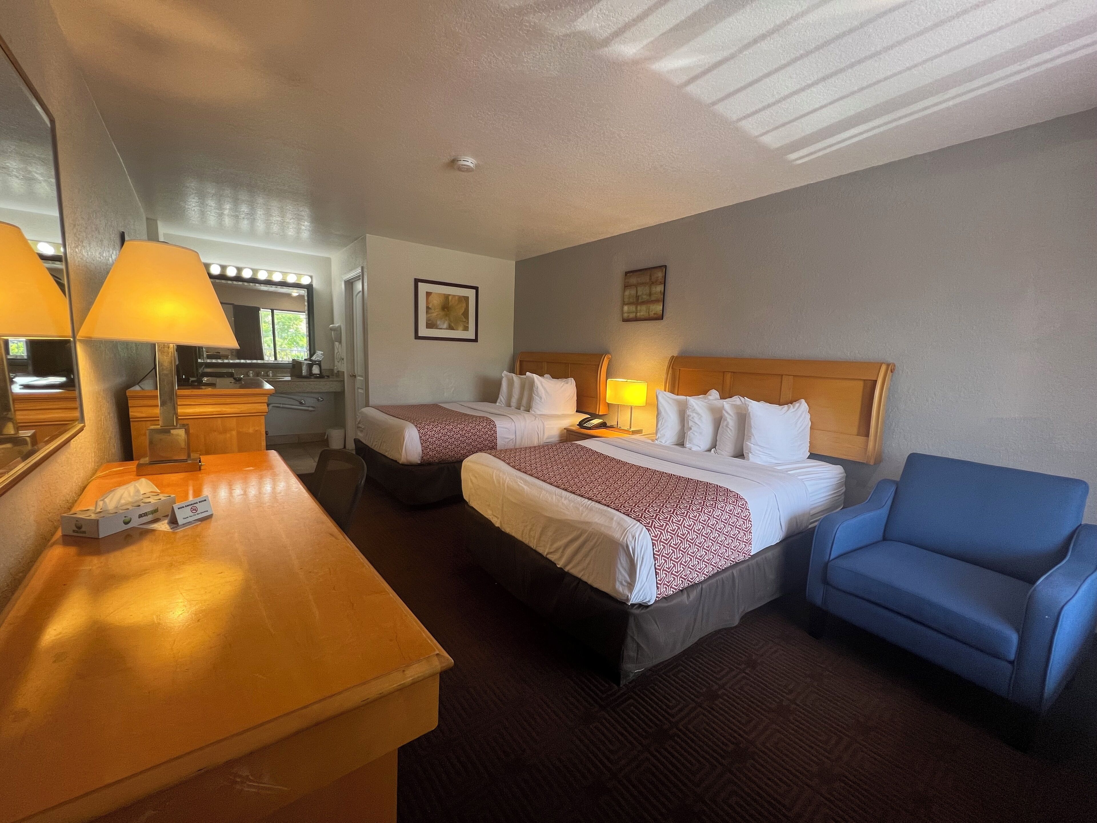 A hotel room with two beds, a desk with a lamp, a chair, a mirror, and a small kitchenette.