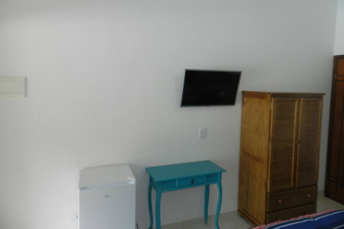 A room with a white wall, a small teal table, a wooden wardrobe, and a black flat-screen TV mounted on the wall.