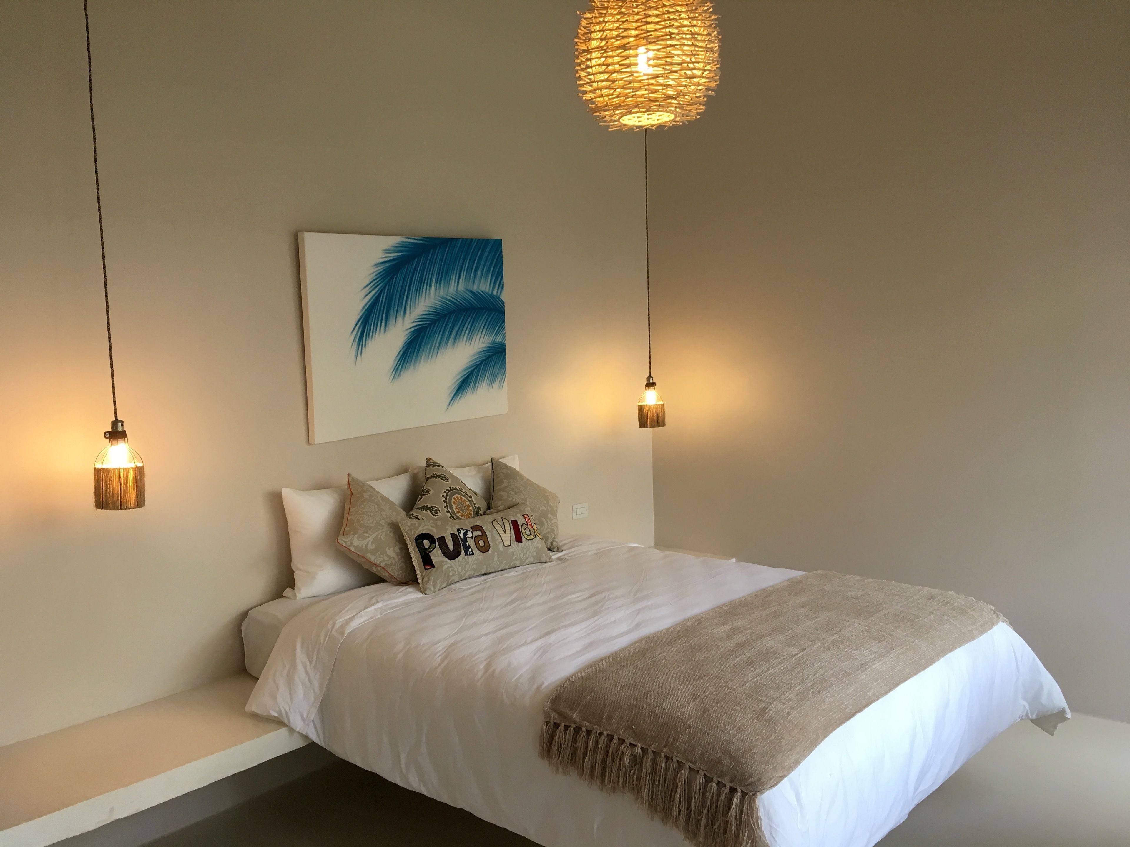 Un dormitorio con una cama, dos lámparas colgantes, una obra de arte en la pared y una manta texturizada.
