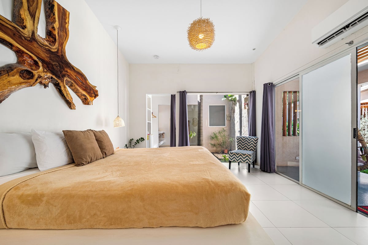 Een moderne slaapkamer met een groot bed, een houten kunstwerk aan de muur, en uitzicht op het groen door een schuifdeur.