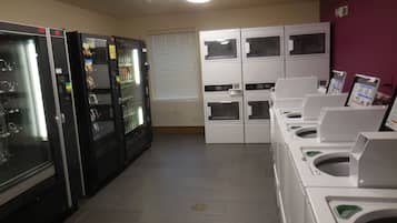A laundromat with multiple washing machines and dryers, a vending machine, and a window with blinds.