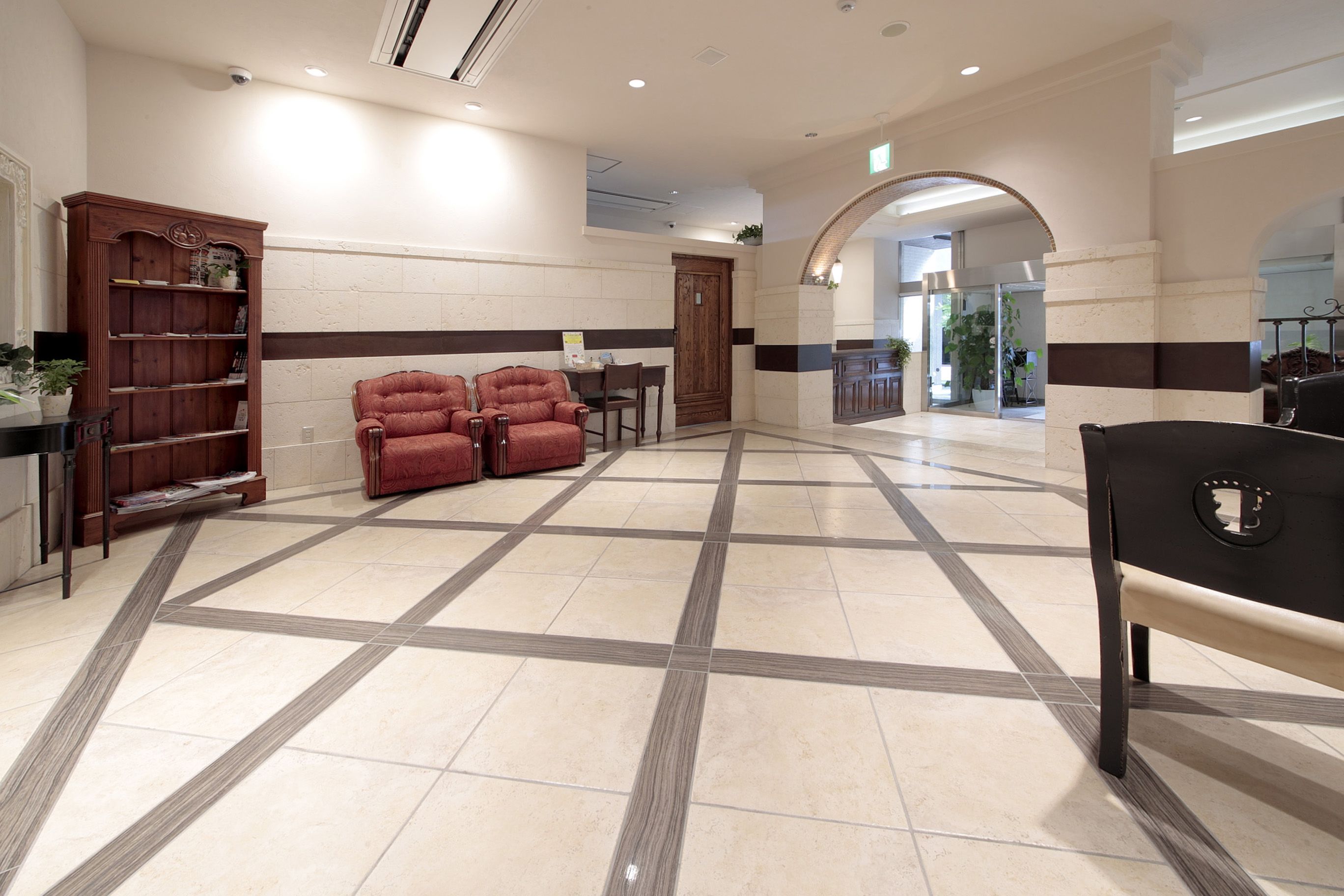 A spacious lobby with a checkered floor pattern, a wooden bookshelf, a red armchair, and a black bench.