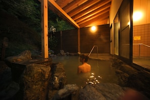 Zona de baño de aguas termales con vigas de madera, rocas y una persona en el agua.