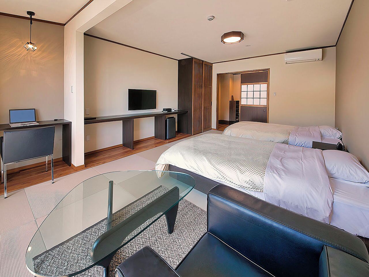 Habitación de hotel moderna con una mesa de centro de vidrio, una cama con ropa blanca, un escritorio con una computadora y un televisor.